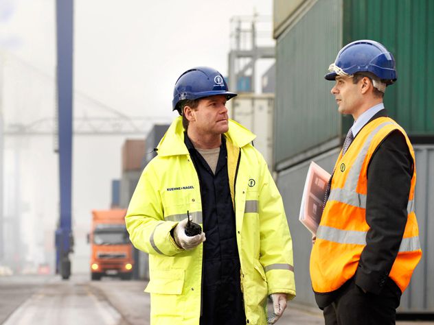 two man talking in front of containers with a bridge in the background